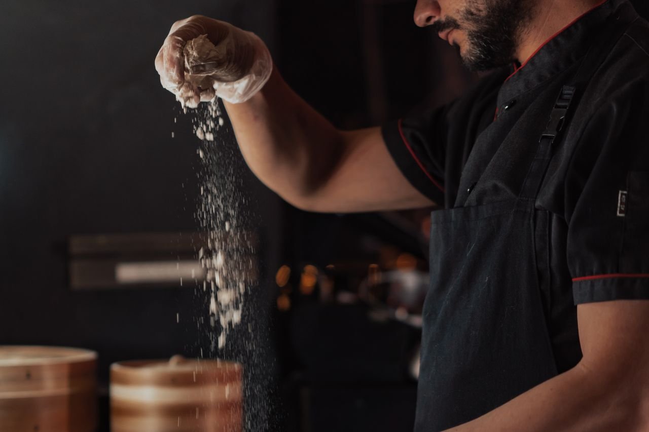 Mastering the First Impression: Your intriguing post title goes here A chef wearing a black apron sprinkles flour onto a surface in a dimly lit kitchen, showcasing food preparation.